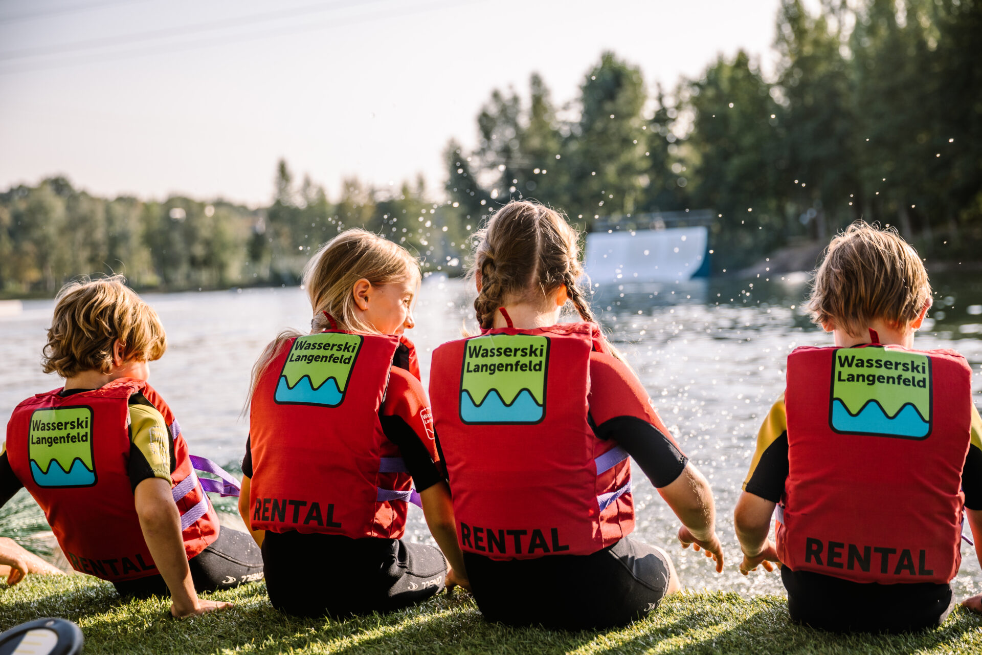 Vier Kinder sitzen mit roter Langenfeled-Schwimmweste, mit dem Rücken zur Kamera, auf dem Startplatz und plantschen mit den Füßen im Wasser.