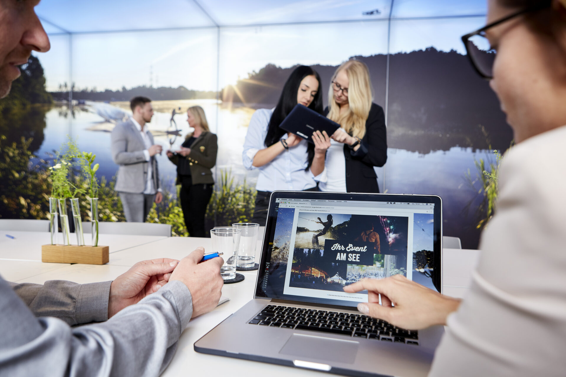 Tagungsraum. Mann und Frau schauen im Vordergrund auf einen Laptop, im Hintergrund besprechen sich ebenfalls zwei Teams