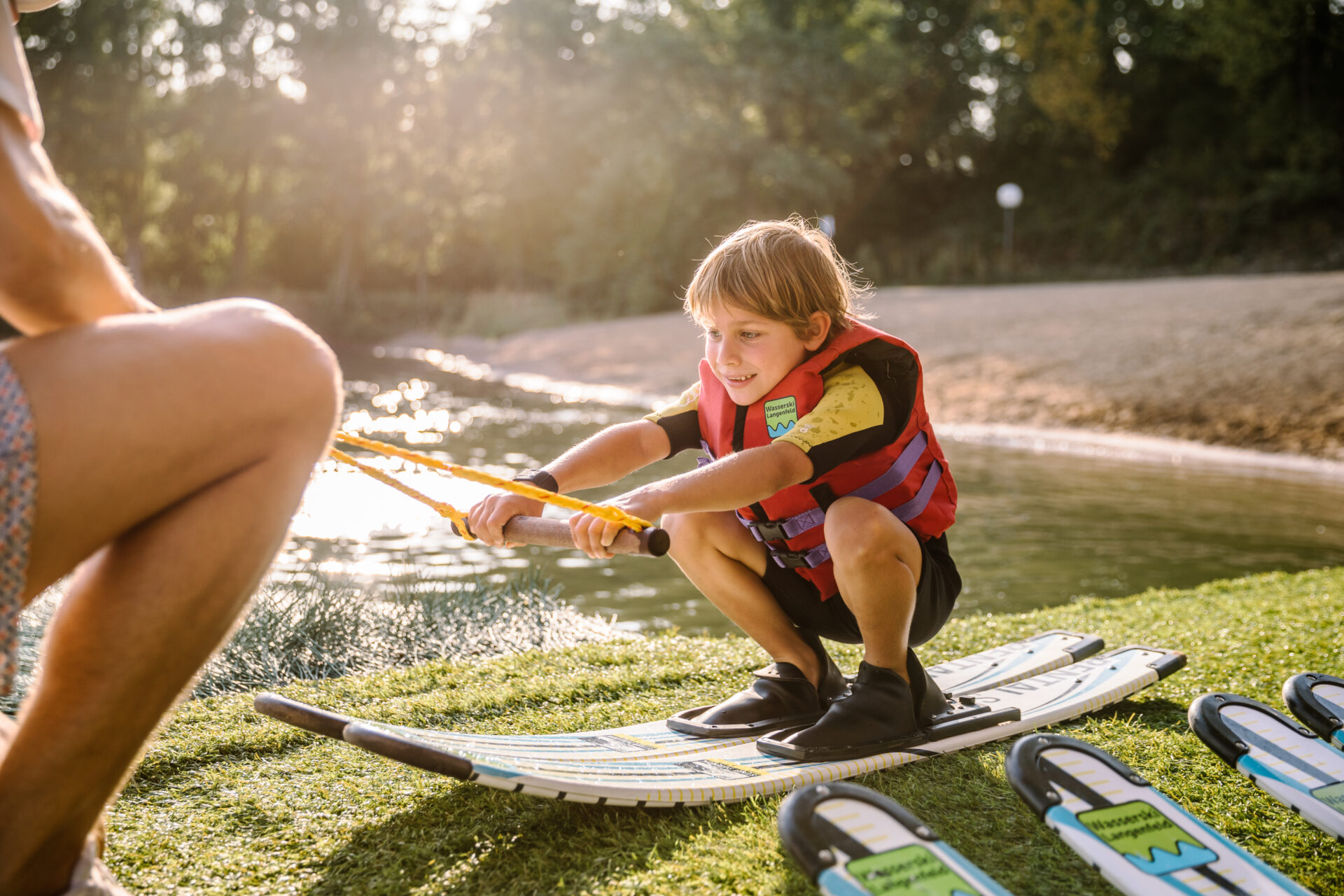 wasserski-langenfeld-minibahn-anfängereinweisung-kind Kleiner Junge sitzt in der Hocke und macht Wasserski-Trockenübung zusammen mit einem Trainer