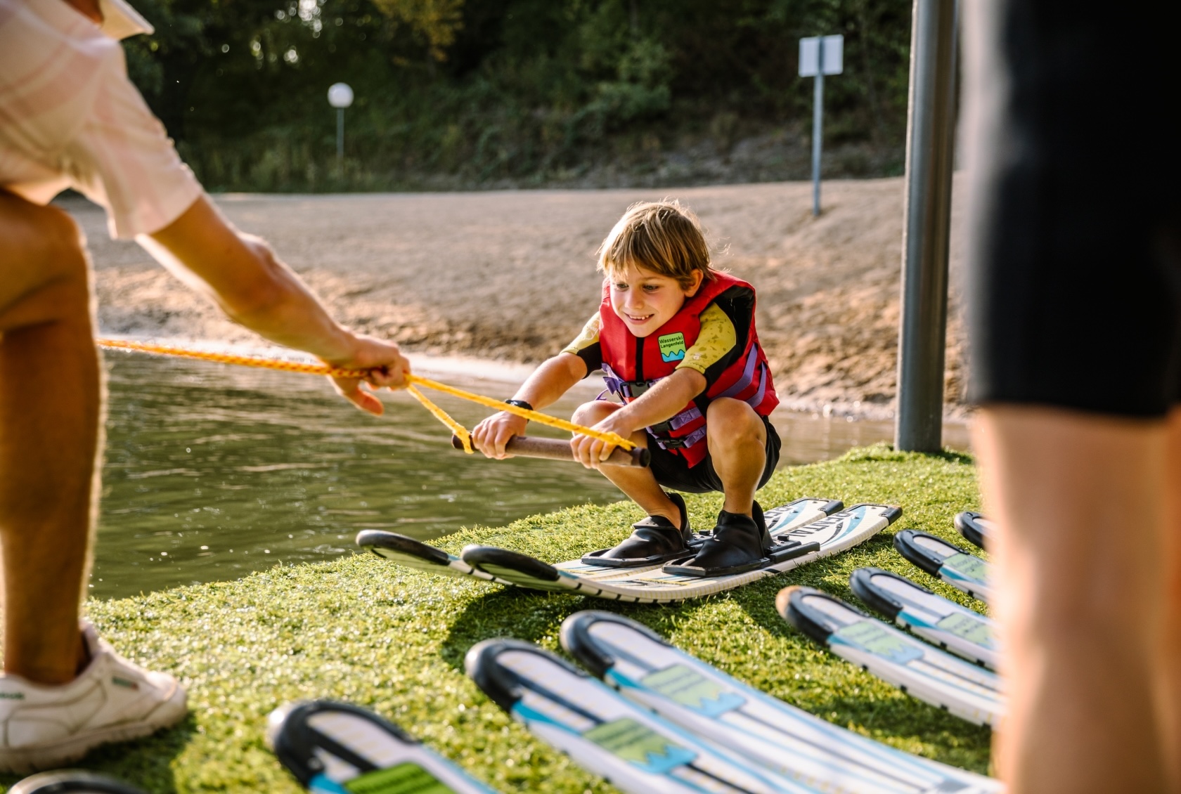 Junge mit roter Schwimmweste und Wasserski an den Füßen in der Hocke. Trockenübung mit Hantel